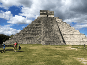 A large stepped pyramid stands under a partly cloudy sky. Two adults and two children walk across a grassy area in front of the structure, which showcases ancient stone steps and intricate design.
