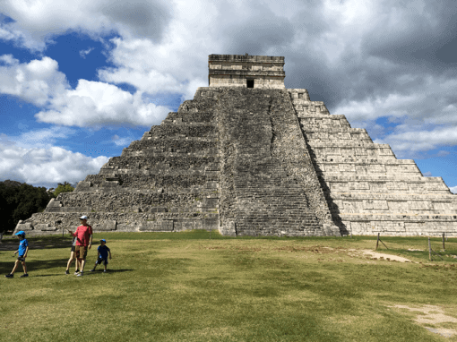 A large stepped pyramid stands under a partly cloudy sky. Two adults and two children walk across a grassy area in front of the structure, which showcases ancient stone steps and intricate design.