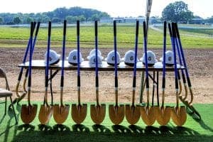 A table displays multiple gold shovels and white hard hats with a logo, set against a grassy field and blue sky.