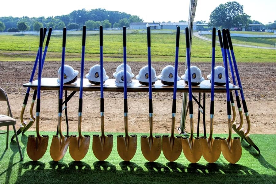 A table displays multiple gold shovels and white hard hats with a logo, set against a grassy field and blue sky.
