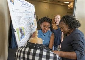 Three people engage around a mannequin display in front of a research poster. One woman with glasses and a denim shirt talks while another woman observes, and a third examines the garment on the mannequin. Background elements include a chandelier and a table.