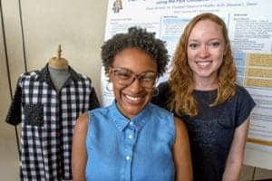 Two women pose together, smiling in front of a research poster. One wears a denim shirt and glasses, while the other has red hair and wears a dark shirt. To the left, there’s a mannequin displaying a black and white checkered shirt.