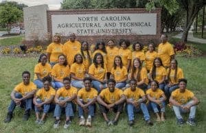 A diverse group of students wearing matching yellow shirts poses in front of a sign for North Carolina Agricultural and Technical State University. They sit on the grass, surrounded by flowers, and display a sense of unity and camaraderie.