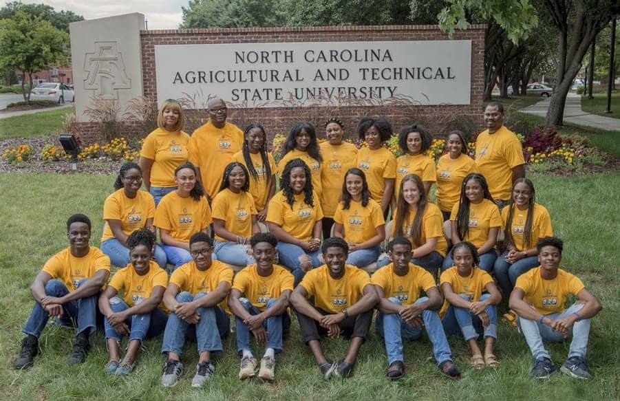 A diverse group of students wearing matching yellow shirts poses in front of a sign for North Carolina Agricultural and Technical State University. They sit on the grass, surrounded by flowers, and display a sense of unity and camaraderie.