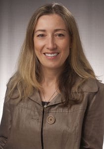 A professional woman with long blonde hair smiling, wearing a brown jacket, in a neutral background.