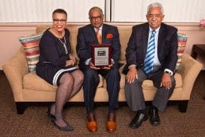 Three people sitting on a couch. The person in the middle holds a plaque. They are all smiling and dressed formally, with the left person wearing a dress, the middle a suit with an orange pocket square, and the right a suit with a blue striped tie.