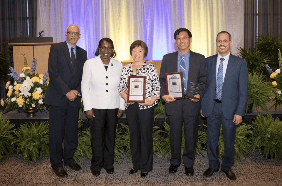 Award ceremony with five professionals holding plaques at a formal event, with floral arrangements a.