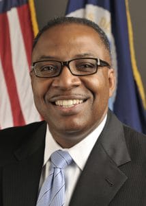Professional African American man in suit smiling with flags in background.