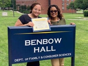 Two women celebrating graduation at Benbow Hall, holding a diploma and smiling.