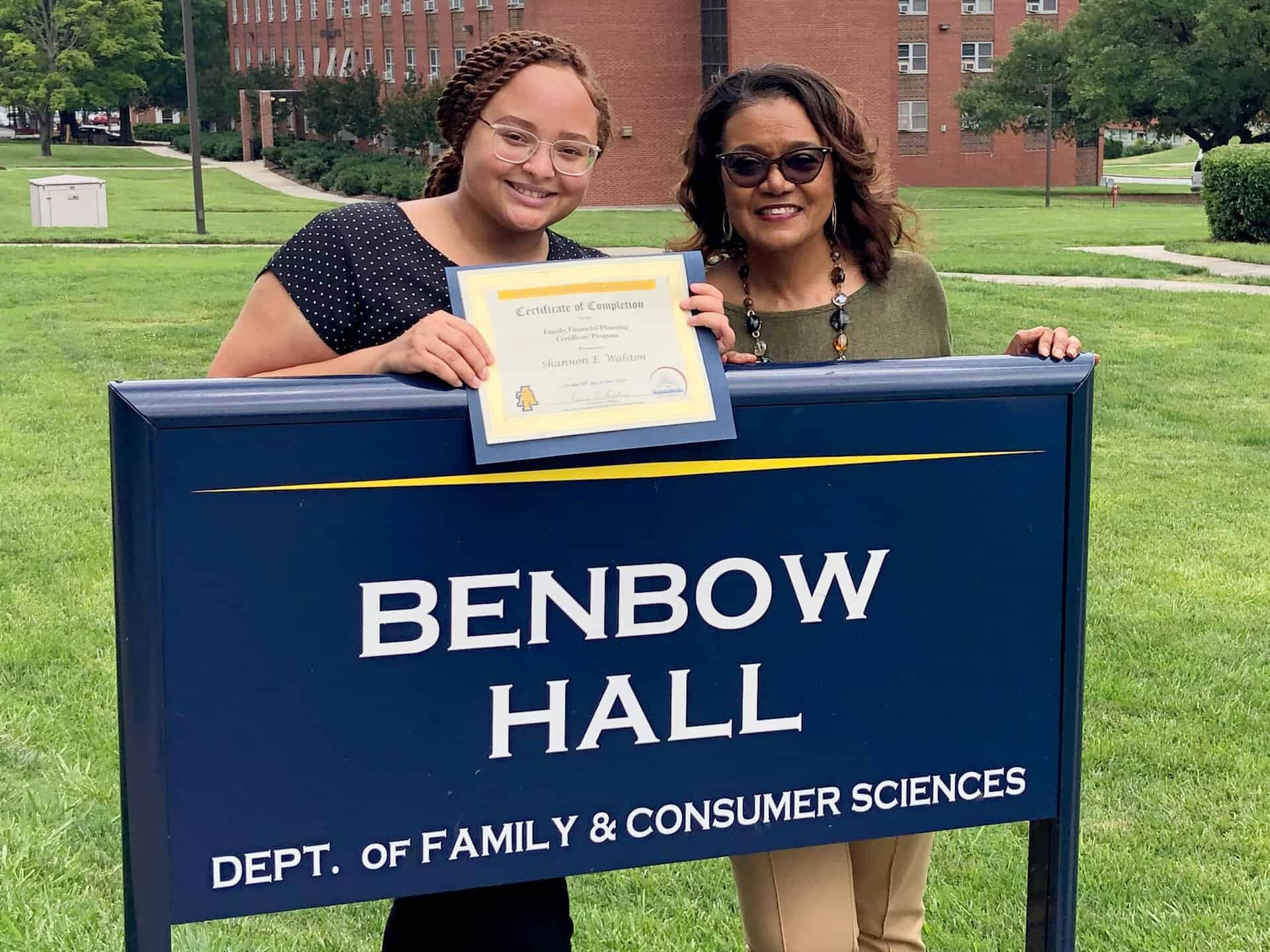 Two women celebrating graduation at Benbow Hall, holding a diploma and smiling.