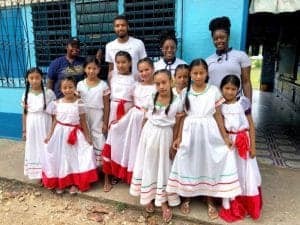 Group of children and adults in traditional white and red dresses outdoors.