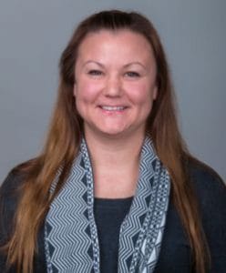 A woman with long brown hair smiling, wearing a patterned scarf and dark blazer.