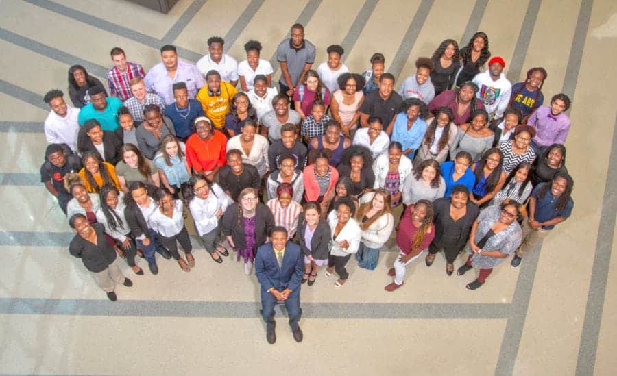A diverse group of students stands together, smiling, in a spacious indoor setting with geometric floor patterns.
