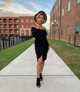 Elegant woman in black dress and hat standing on sidewalk in urban residential area at sunset.
