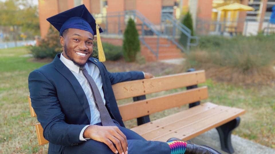 Graduating student in cap and gown sitting on park bench outdoors.