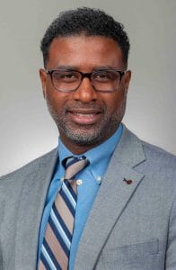 Professional man in a gray suit and glasses, smiling confidently for a corporate headshot.