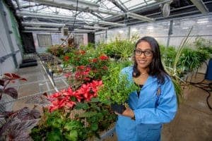 Woman in a blue lab coat holding a potted plant in a greenhouse with lush greenery.
