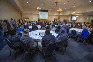 Conference attendees listening to a speaker at a professional event in a large meeting room.