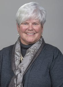 A woman with short white hair smiling, wearing a dark gray top and a patterned scarf.