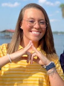 A woman smiling outdoors near water, wearing glasses and a yellow striped shirt.