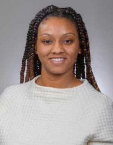 A woman with braided hair smiling at the camera, wearing a light-colored sweater.