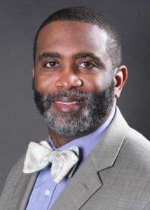 A man in a light gray suit with a bow tie, smiling confidently against a dark background.