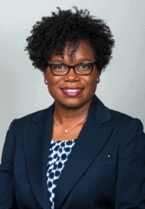 A woman with curly black hair and glasses wearing a navy blazer and patterned blouse.