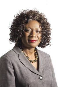 Elegant African American woman in professional gray blazer with curly hair and pearl necklace.