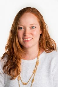 A woman with red hair smiling, wearing a white top and gold necklace, against a plain white backgrou.