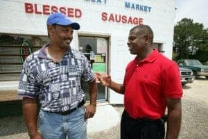 Two men smiling and talking outside a store with "Blessed Sausage" sign.