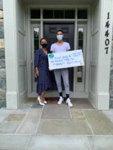 A woman and a man holding a large check outside a building, wearing masks.