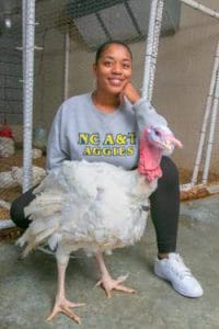 Happy young woman with a turkey in a farm setting, smiling and wearing a gray sweatshirt.