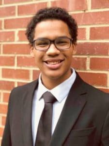 A young man in a suit smiling in front of a brick wall.