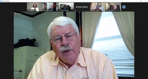 Senior man speaking during a virtual meeting on a computer screen, with multiple participants visibl.