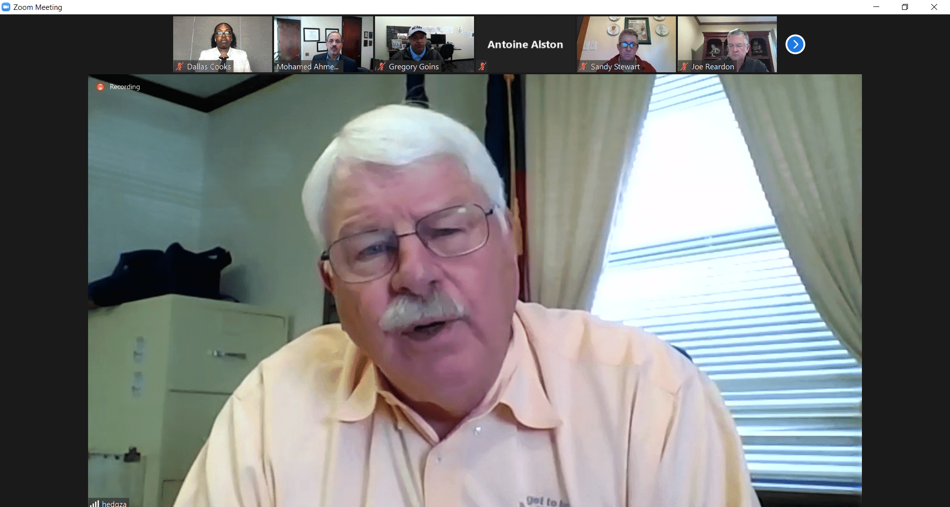 Senior man speaking during a virtual meeting on a computer screen, with multiple participants visibl.