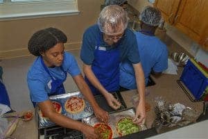 Preparing homemade pizzas in a kitchen with a diverse group of people wearing blue aprons and hairne.