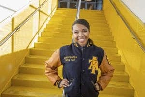 Smiling woman in a varsity jacket stands on bright yellow stairs, showcasing school pride.