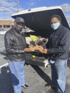 Two men exchanging food outdoors near a car trunk, wearing masks and jackets.