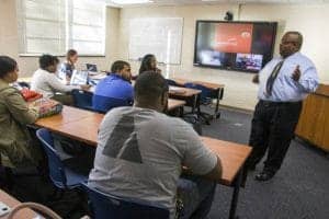 High school students attending a classroom lecture with a teacher presenting at the front.