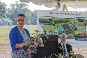 A woman speaking at an outdoor event with a university farm complex map in the background.