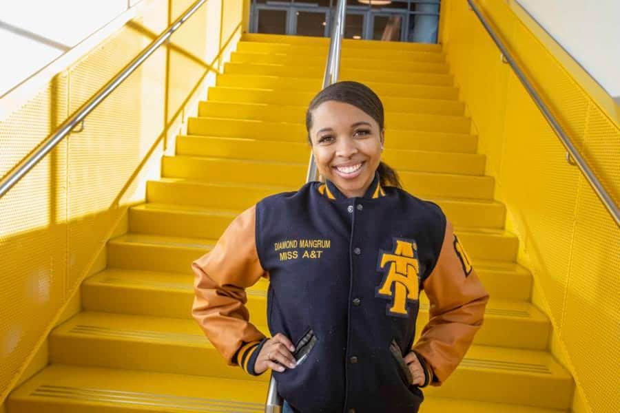 A woman smiling on yellow stadium stairs wearing a black and orange jacket with "Diamond Mangrum Mis.