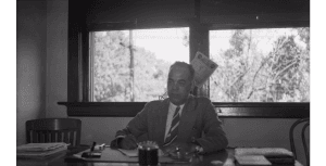 A man in a suit sitting at a desk in an office with a window behind him.