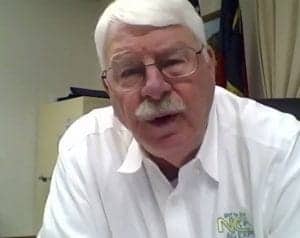 A senior man with white hair and glasses speaking in an office setting.