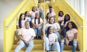 A group of diverse young women sits smiling on yellow stairs, wearing matching T-shirts in pastel colors.