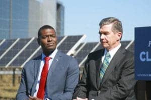 Two men in business suits speaking outdoors at a formal event, with a government sign in the backgro.
