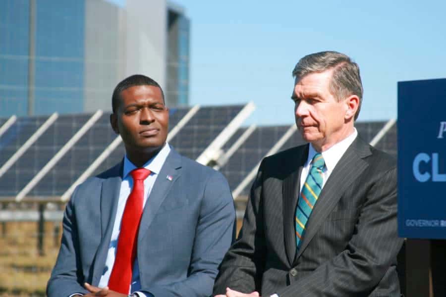 Two men in business suits speaking outdoors at a formal event, with a government sign in the backgro.
