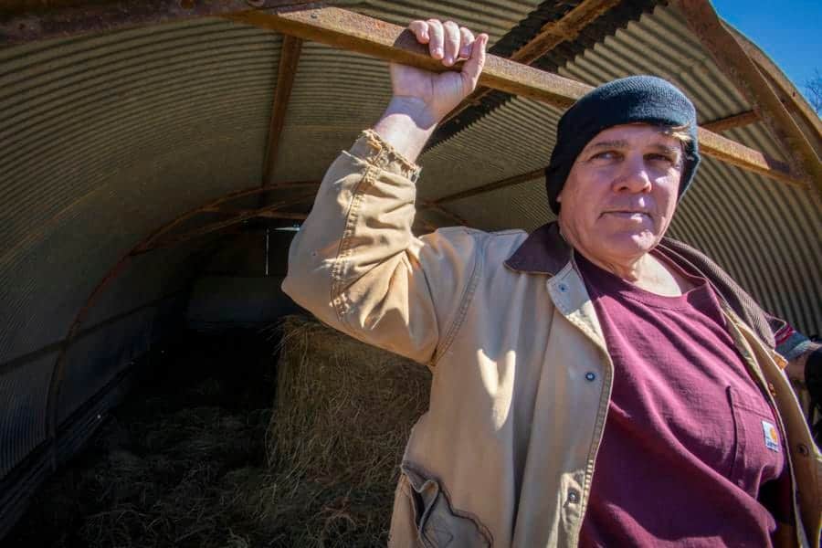 A woman holding a wooden stick inside a barn or shed, wearing a beanie and casual clothing.