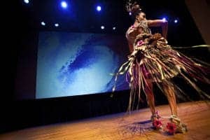 Elegant dancer in traditional costume performing on stage with vibrant lighting and digital backdrop.