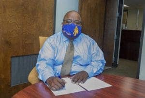 A person wearing a blue mask with yellow letters sits at a wooden table in an office. They are dressed in a light blue shirt and patterned tie, holding a pen over papers.