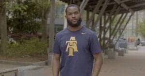 A man standing outdoors wearing a navy blue T-shirt with gold logo, urban background.
